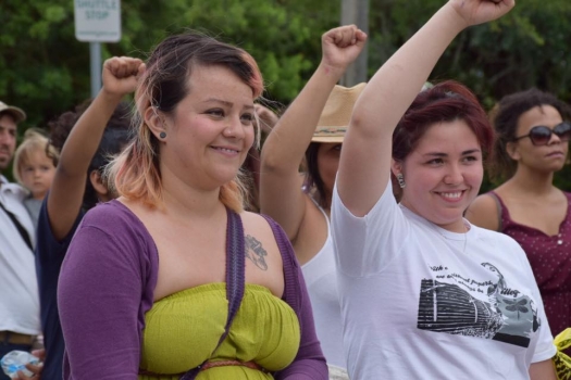 A crowd of people smiling with their fists raised at a demonstration outside the Boca Raton headquarters of GEO Group, a for-profit prison company
