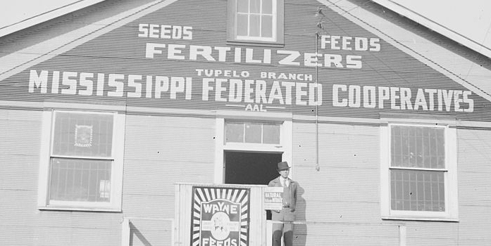 In a black and white photo, a man stands in front of the door of a house featuring a sign reading "Tupelo Branch: Mississippi Federated Cooperatives"